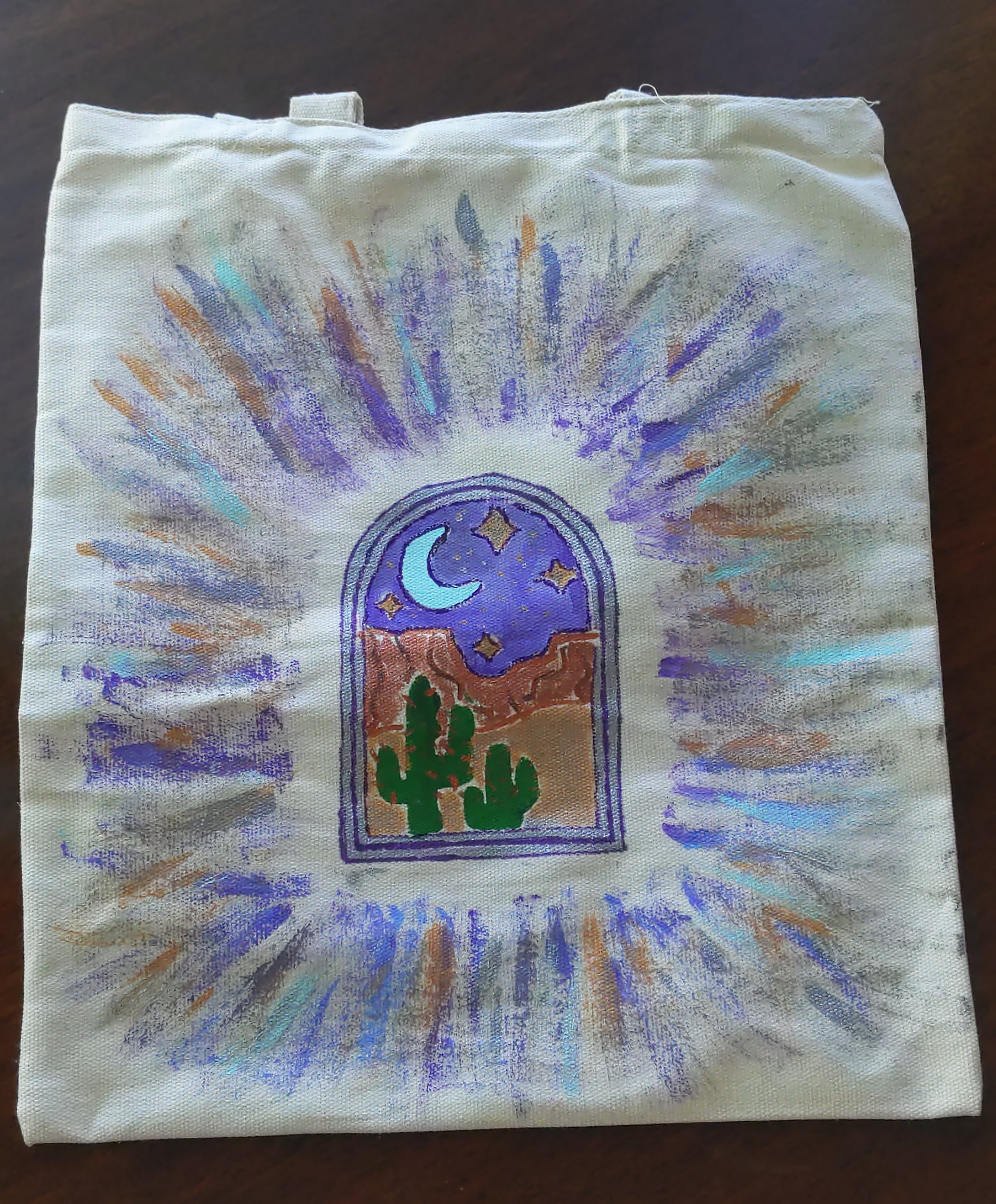 A canvas tote bag with a desert scene painted on it in bright colors